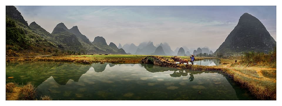 A panoramic view of Guilin's iconic karst mountains reflects perfectly in the still waters, creating a mirror world of ethereal beauty. The lone figure tending to water buffaloes adds a touch of human scale to this vast, tranquil landscape.