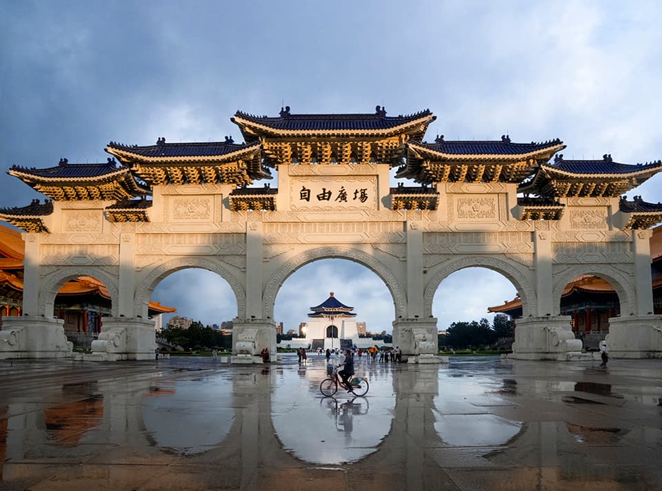 The ornate entrance to Taipei's Chiang Kai-shek Memorial Hall glows in the fading light, its grand architecture a testament to cultural heritage. The rain-slicked ground creates a mirror effect, doubling the visual impact of this impressive structure.