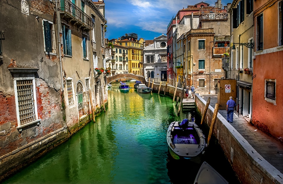 A vibrant Venetian canal lined with colorful, weathered buildings. The emerald water reflects the architecture, while boats and pedestrians bring life to this iconic Italian cityscape, embodying centuries of history and culture.