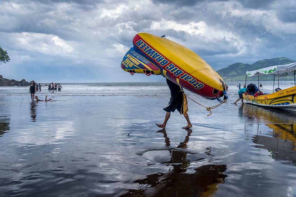 A beach vendor carries a large, colorful inflatable raft across wet sand, set against a backdrop of stormy skies and green hills. This image captures the resilience and entrepreneurial spirit of coastal communities, even in challenging weather.