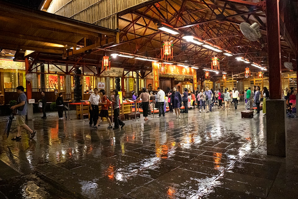 A bustling night market under a traditional wooden structure, its rain-slicked floor reflecting the warm glow of lanterns. Visitors mingle and explore the stalls, creating a lively atmosphere that contrasts with the dark, wet night outside.