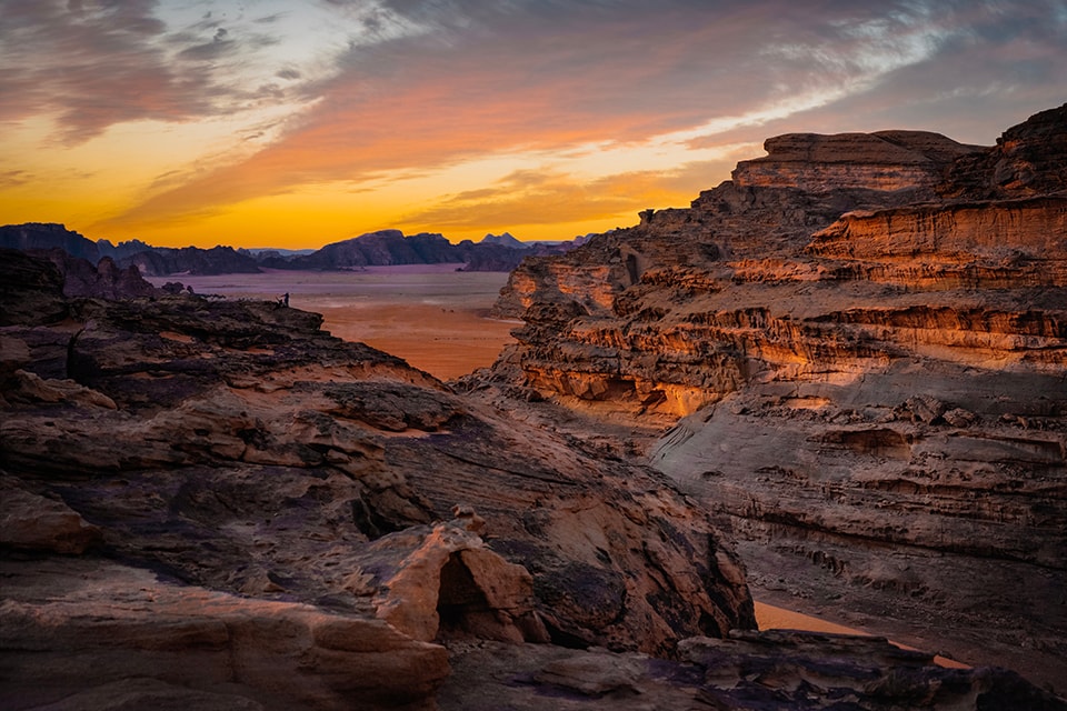 A breathtaking sunset over Wadi Rum, Jordan, paints the sky in vibrant oranges and golds. The layered, eroded rock formations create a dramatic foreground, their warm tones echoing the sky. A tiny human figure on a distant outcrop provides scale, emphasizing the vast, otherworldly nature of this desert landscape.