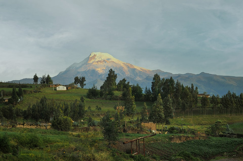 A majestic snow-capped mountain dominates the skyline, its peak catching the golden light. The foreground showcases a lush, terraced landscape dotted with trees and small buildings, exemplifying the harmonious coexistence of nature and human settlement in the Andean highlands.
