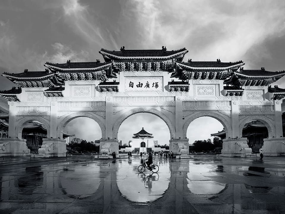 A majestic archway stands as a testament to both tradition and progress. The intricate details of the Chinese architecture contrast beautifully with the modern figure on a bicycle, creating a powerful juxtaposition of old and new. The rain-slicked ground mirrors the grand structure, doubling its impact and symbolizing reflection on the past while moving forward.