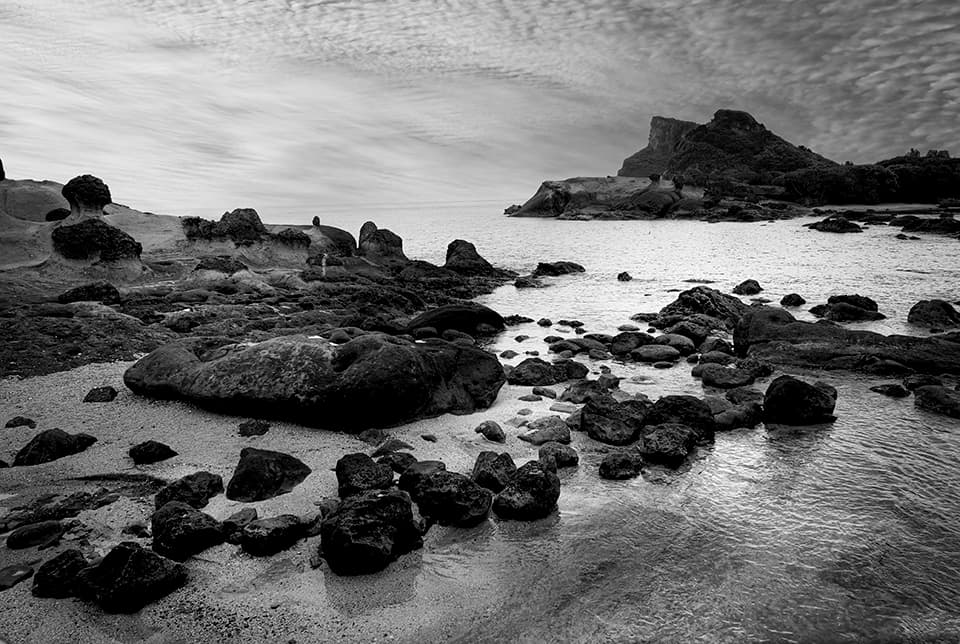 A rugged coastline unfolds before us, its rocky sentinels standing guard against the relentless sea. The interplay of textures - from smooth, wave-worn boulders to jagged cliff faces - creates a visual symphony in black and white. The distant headland, shrouded in a soft haze, adds an air of mystery and beckons the eye to explore further. This image speaks to the enduring strength of nature and the timeless dialogue between land and sea, inviting contemplation on the forces that shape our world.
