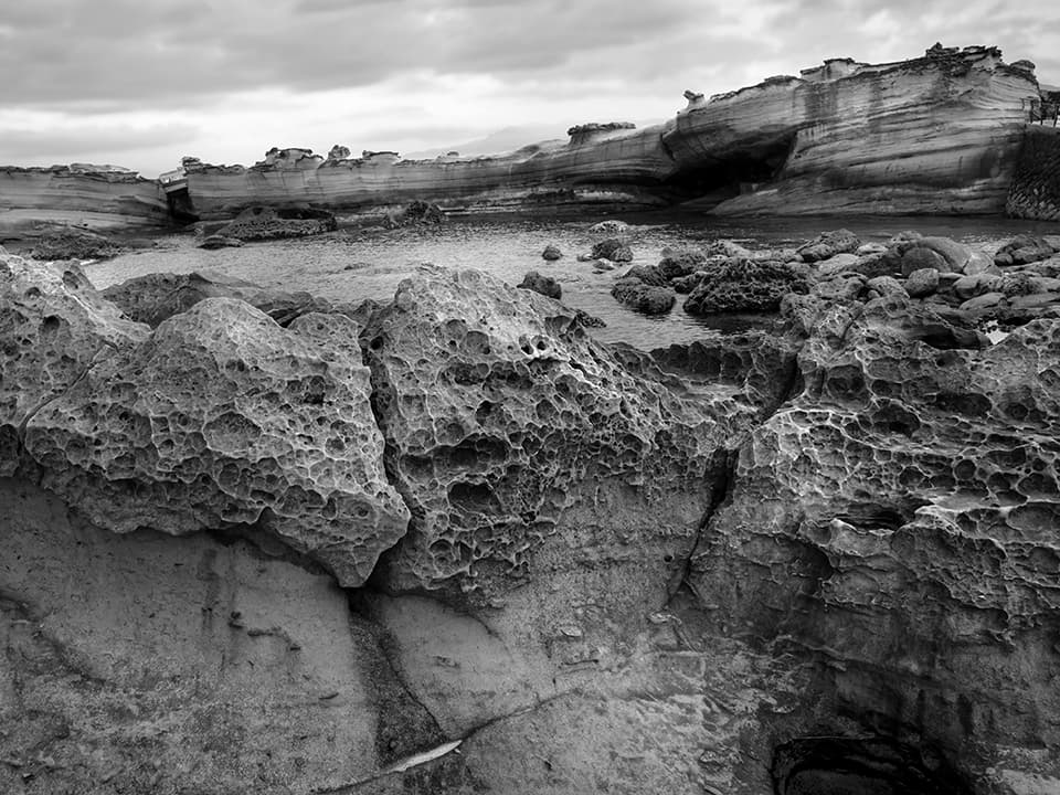 An otherworldly landscape reveals itself in striking detail. In the foreground, intricately eroded rocks resemble a field of alien sculptures, their honeycomb patterns testament to the patient artistry of wind and water. Beyond, layered cliffs rise like ancient fortifications, their strata telling a geological story millions of years in the making. This image celebrates the raw beauty of erosion and time, showcasing nature's ability to craft masterpieces from stone. It invites viewers to marvel at the intricate details and grand scale of our planet's natural wonders.