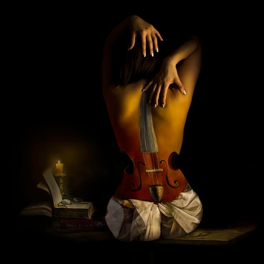 A striking black and white image of a reclining figure whose body is adorned with intricate artwork. The subject's torso features a portrait, possibly of a famous artist, blending human canvas with classical portraiture. This piece challenges perceptions of art and the human form